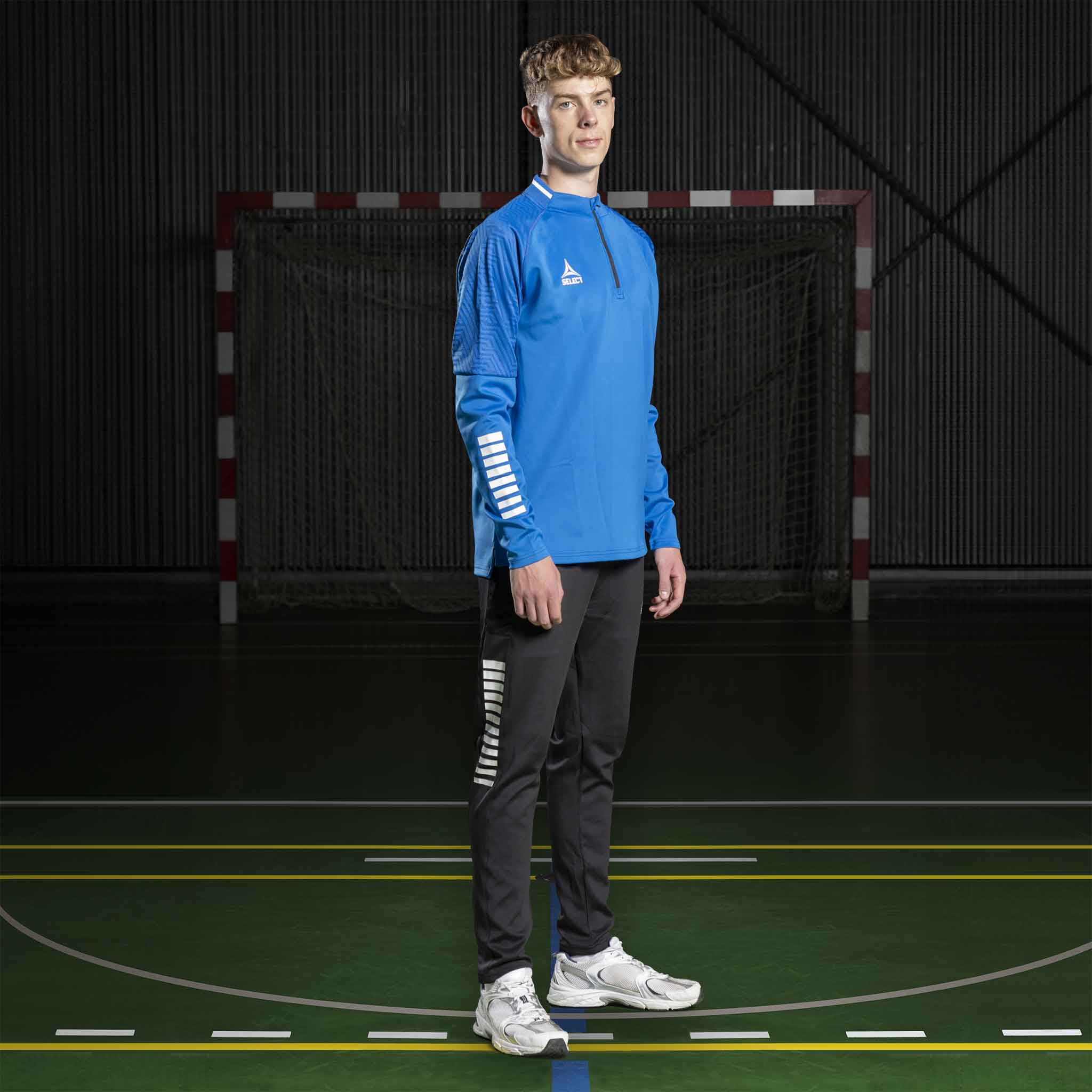Mannlig håndballspiller i blå Training sweat 1/2 zip Monaco v24 og sorte bukser, Match & Training wear, forfra.