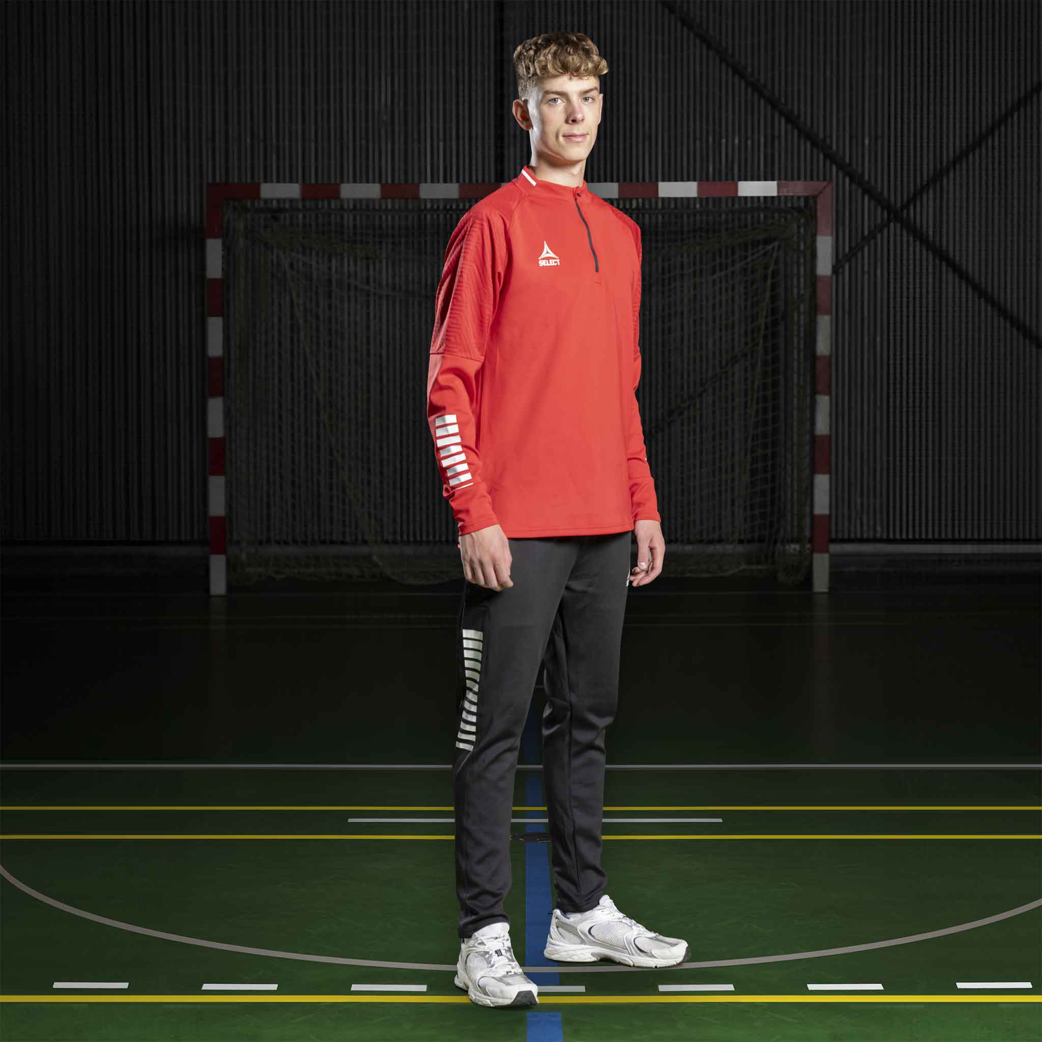 Mannlig håndballspiller i rød Training sweat 1/2 zip Monaco v24 og svarte bukser, Match & Training wear, på bane.