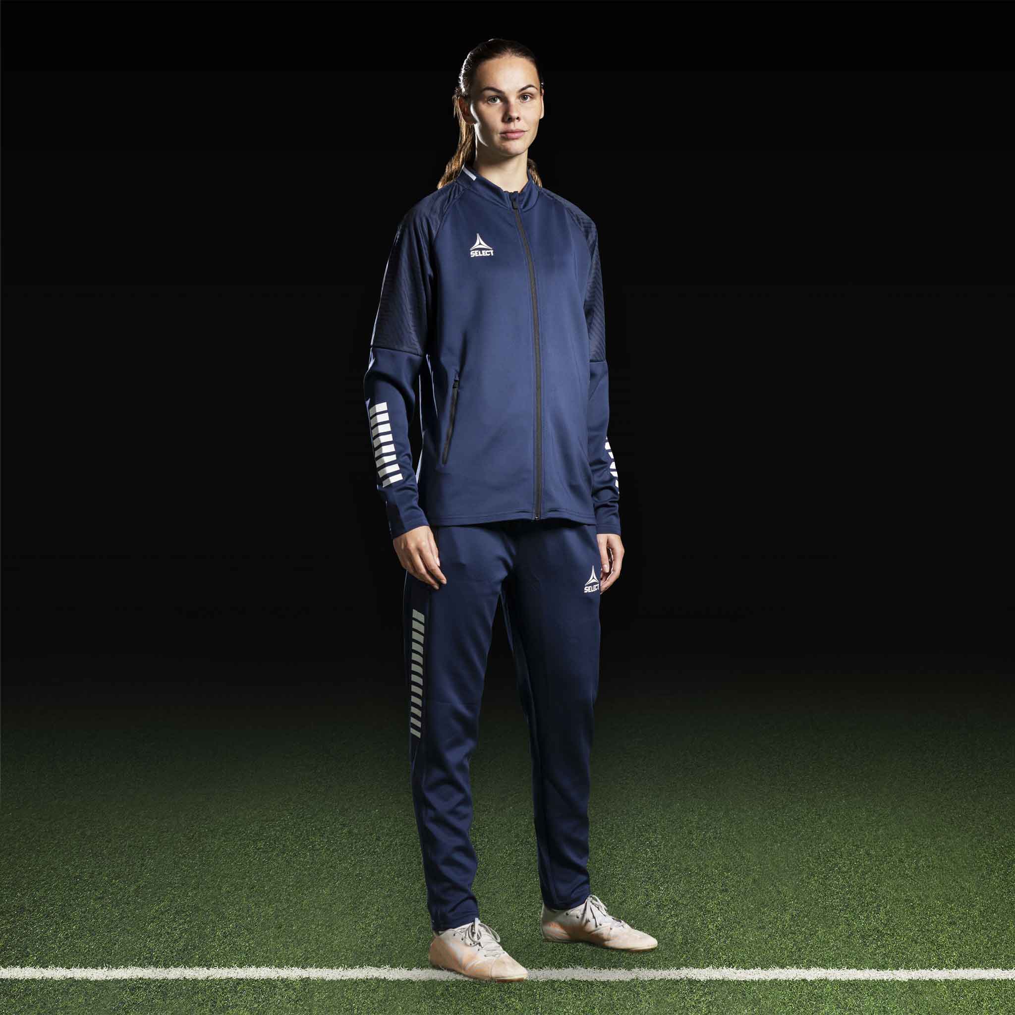 Kvinnelig fotballspiller i marineblå og hvit Zip jacket Monaco v24, Match & Training wear, forfra på banen.