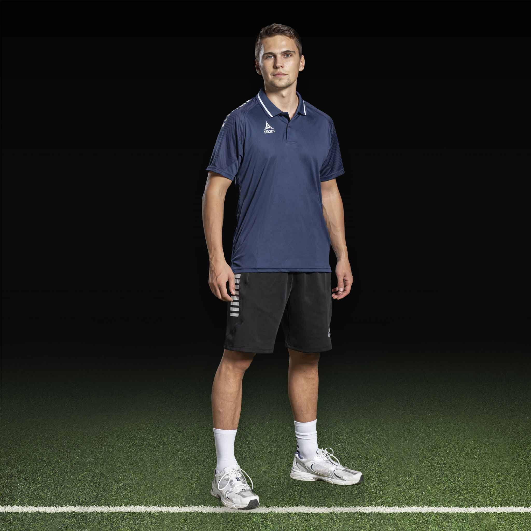 Mannlig fotballspiller i marineblå poloskjorte og sorte Bermuda shorts Monaco v24, leisure wear, forfra på banen.
