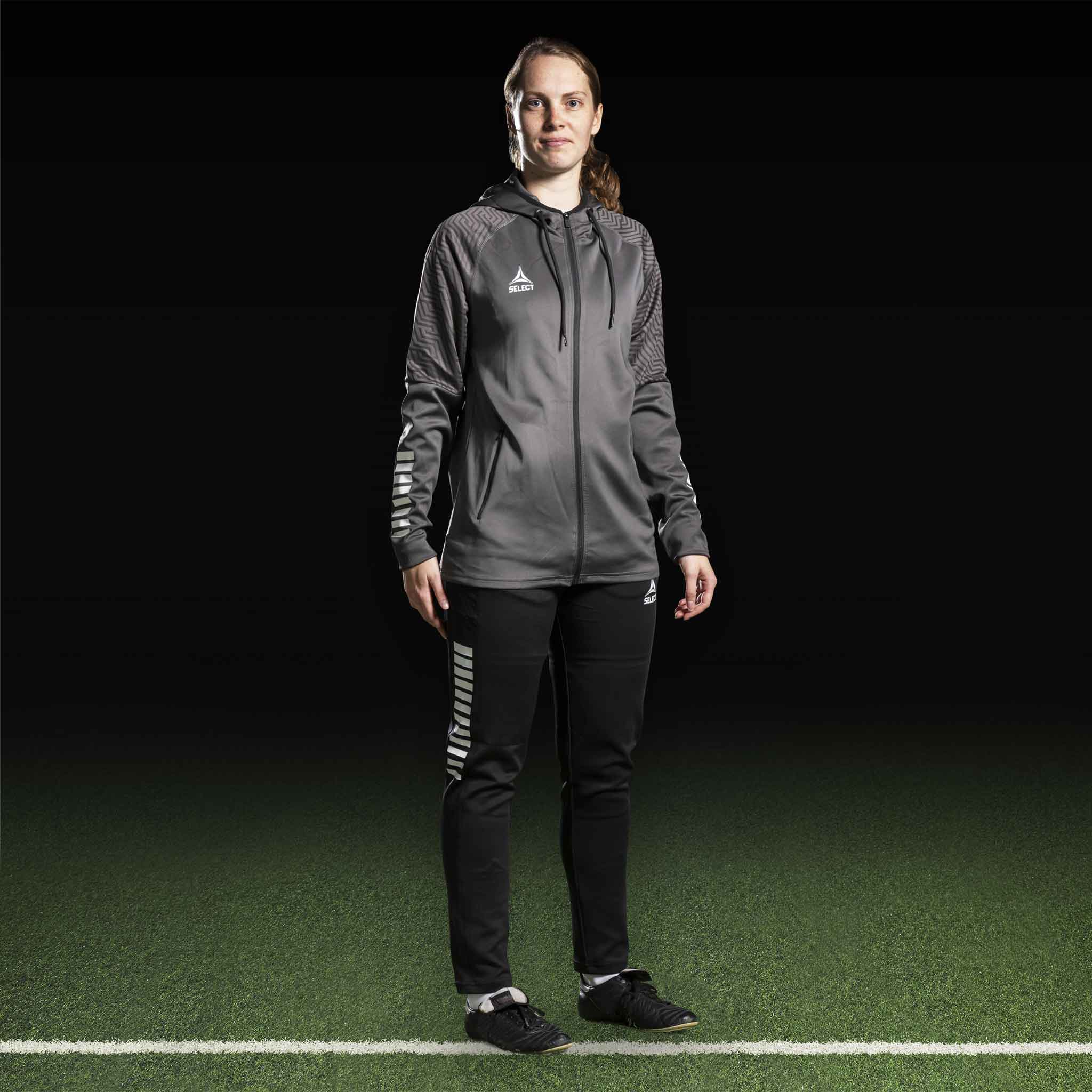 Kvinnelig fotballspiller i grå Zip hoodie Monaco v24 og svarte bukser, Match & Training-tøy.