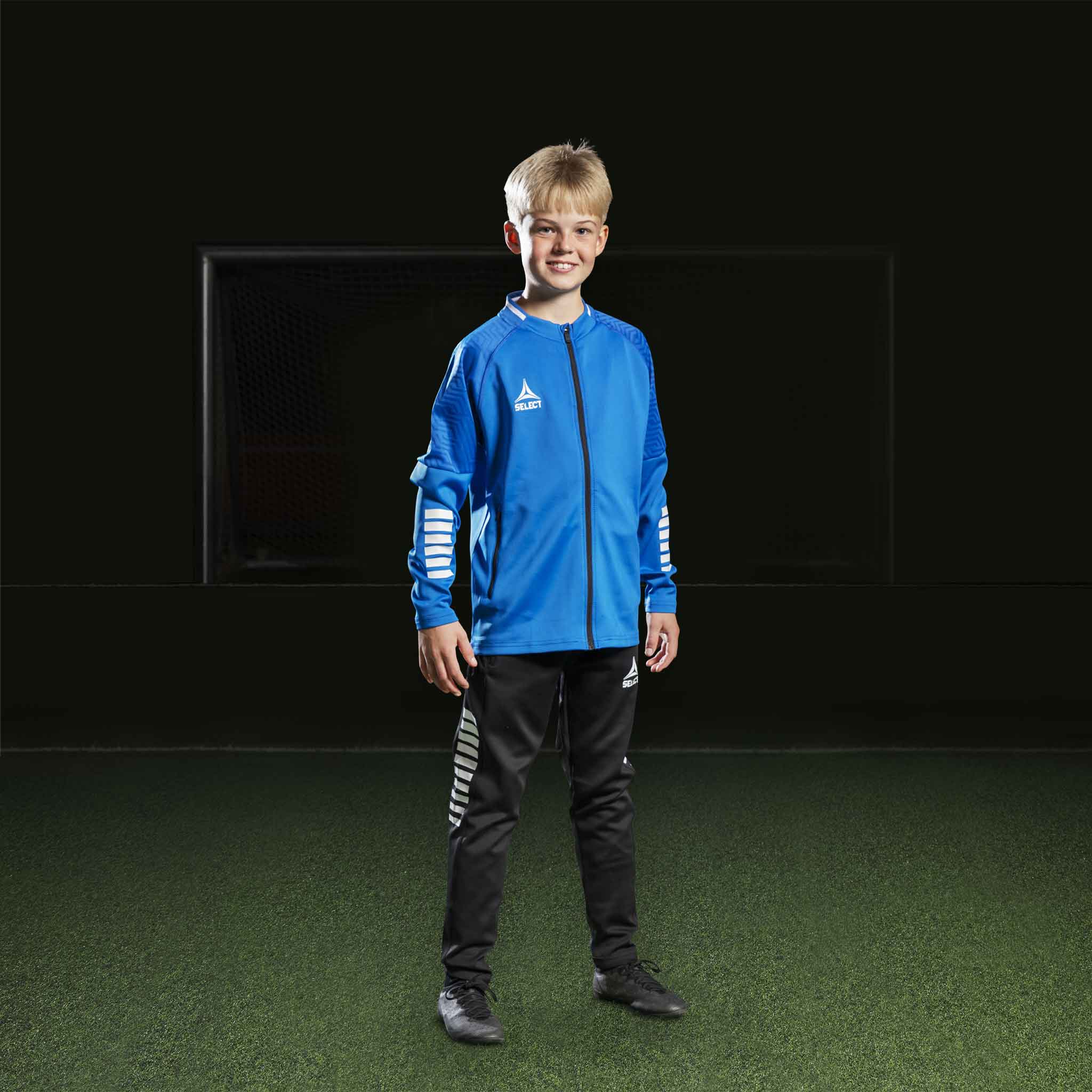 Gutt fotballspiller i blå/hvit Zip jacket Monaco v24 og sorte bukser, Match & Training wear, forfra.