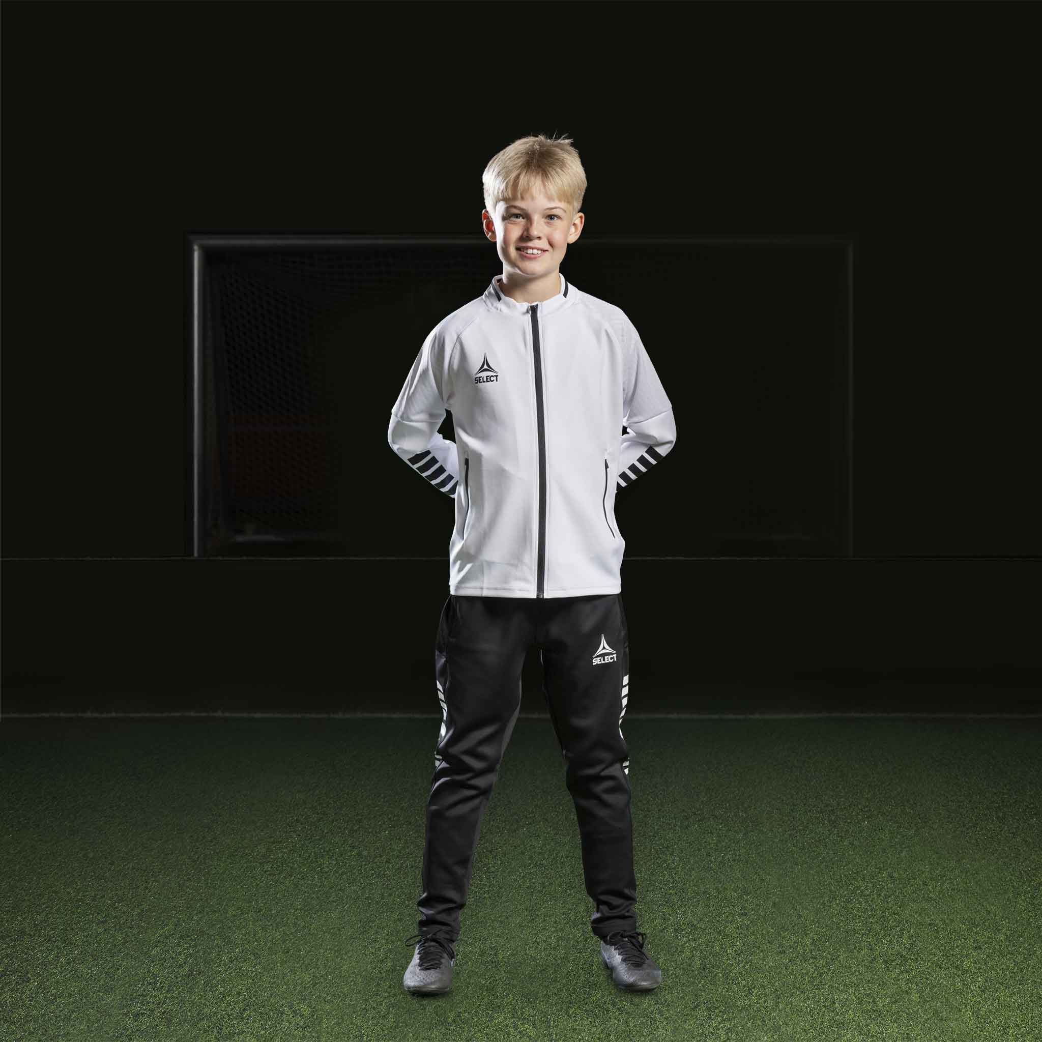 Gutt fotballspiller i hvit Zip jacket Monaco v24 og sorte bukser, Match & Training wear, sett forfra.