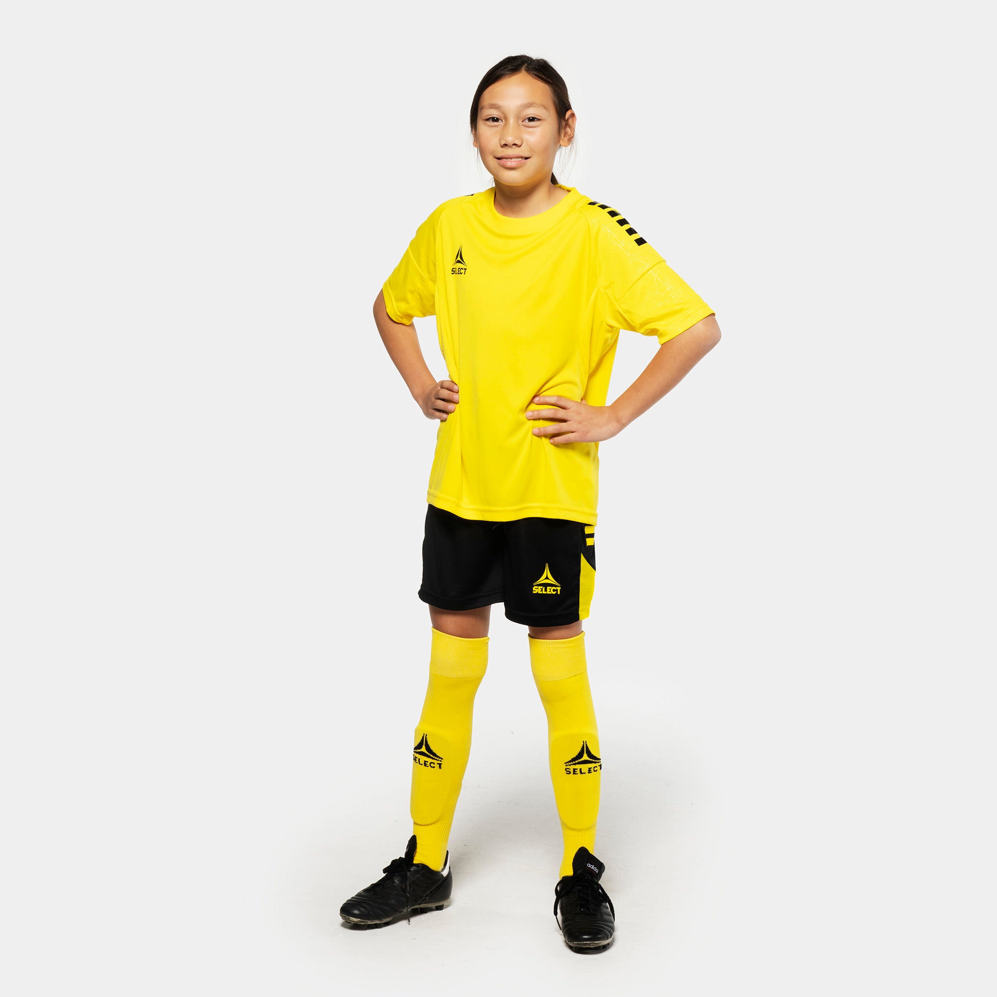 Jente fotballspiller i gul trøye, sorte Player shorts Spain v25 og gule strømper. Match & Training wear, forfra.