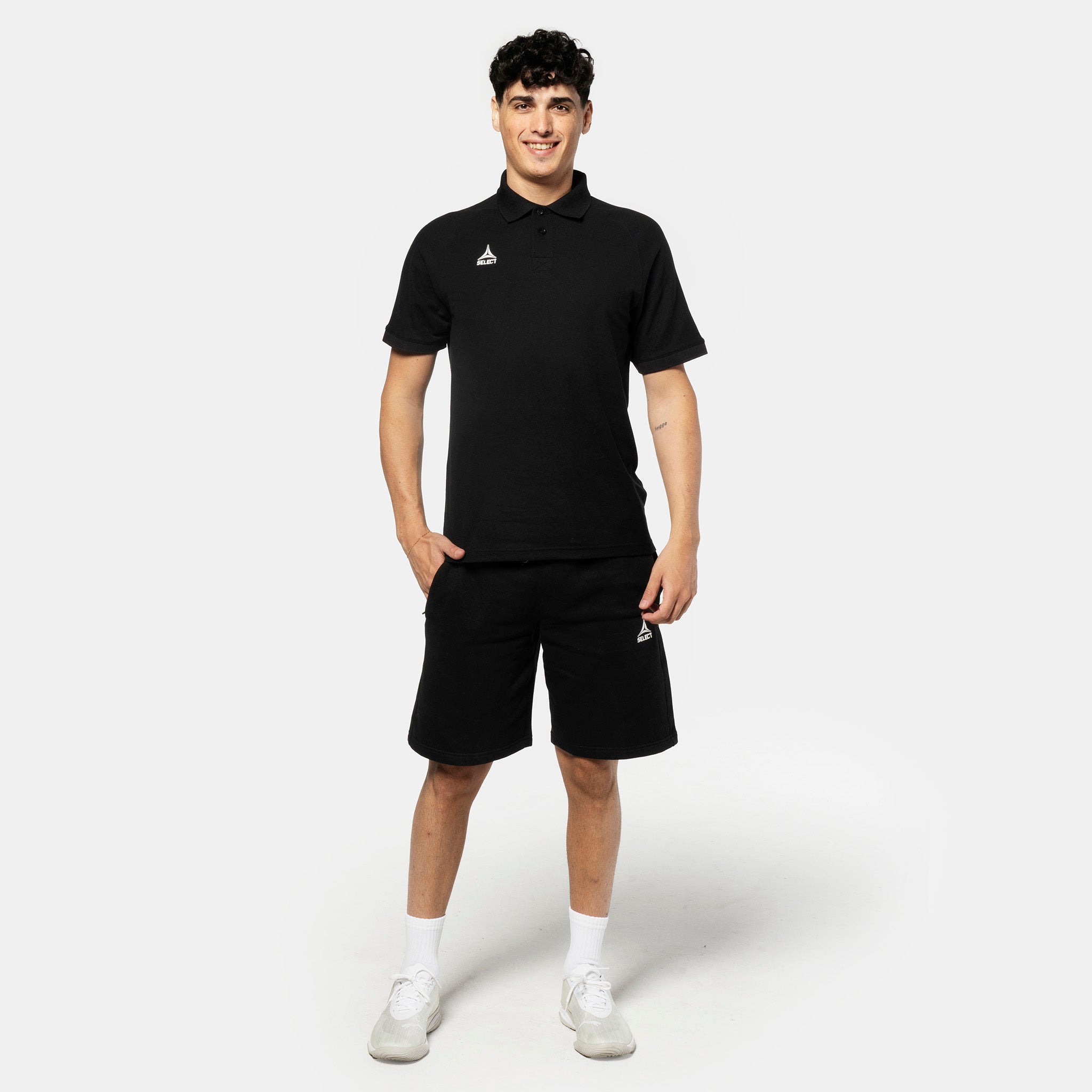 Mannlig fotballspiller i sorte Sweat shorts Oxford v25 fritidsklær, sett forfra med matchende poloskjorte og hvite sneakers.