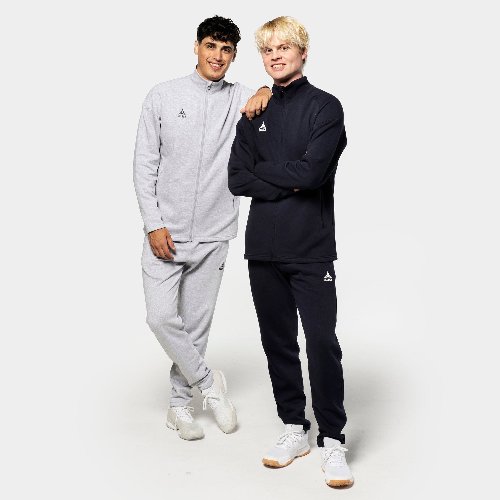 To mannlige fotballspillere i fritidsklær viser Sweat pants Oxford v25 i grått og sort med matchende glidelåsjakker.