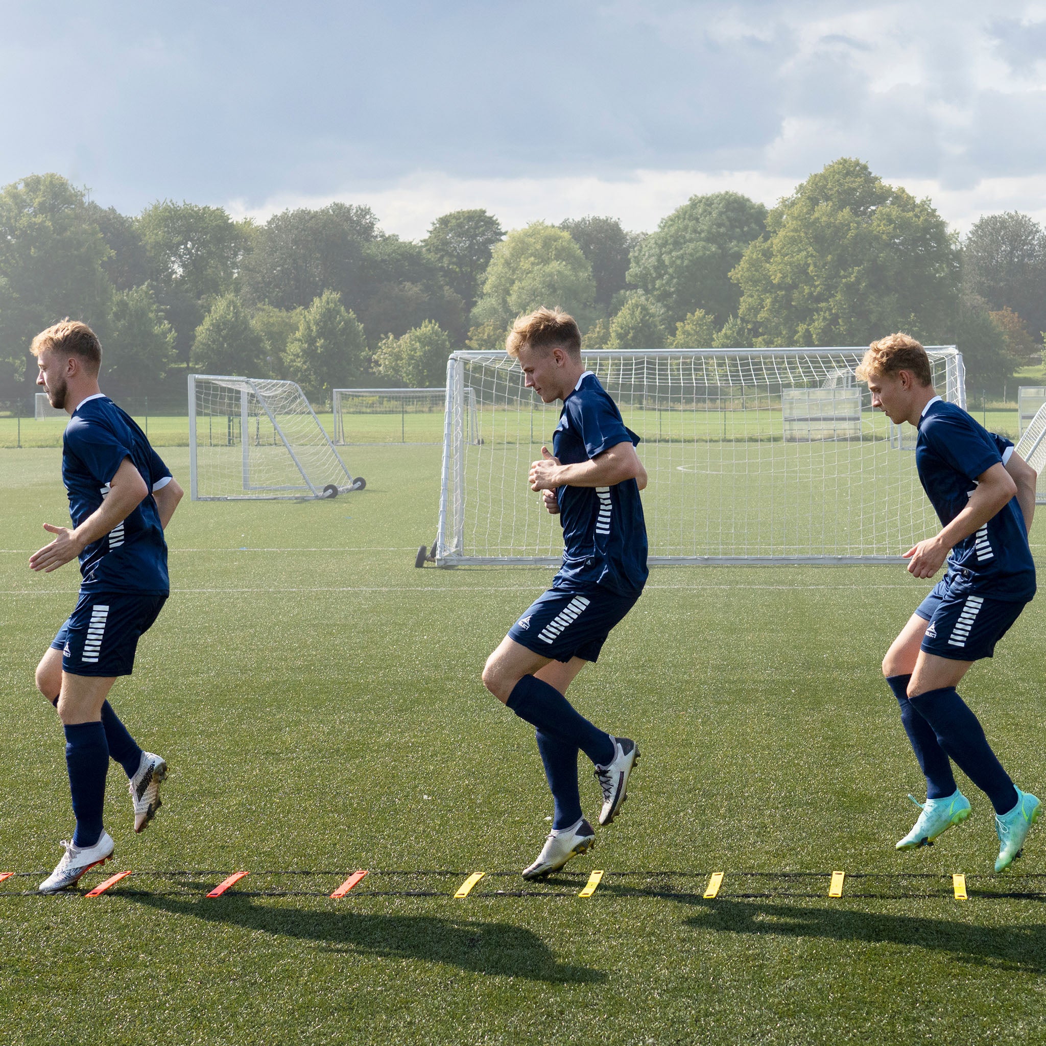 Fotballspillere trener med oransje og gul Agility ladder treningsutstyr på gressbane.