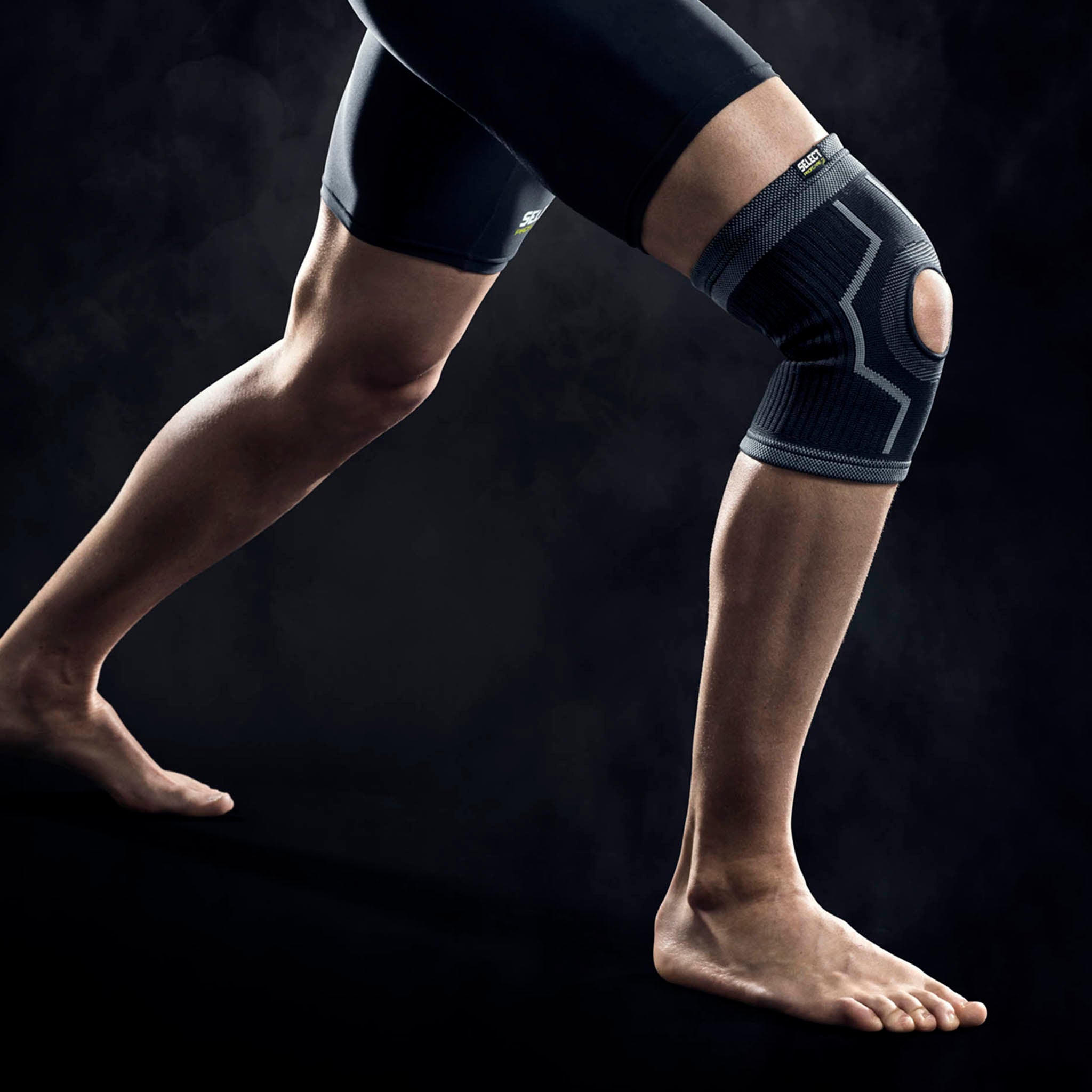 Mann med svart Knee support w/hole, en sportsstøtte, på kneet. Modellfoto som viser forsiden av støtten.